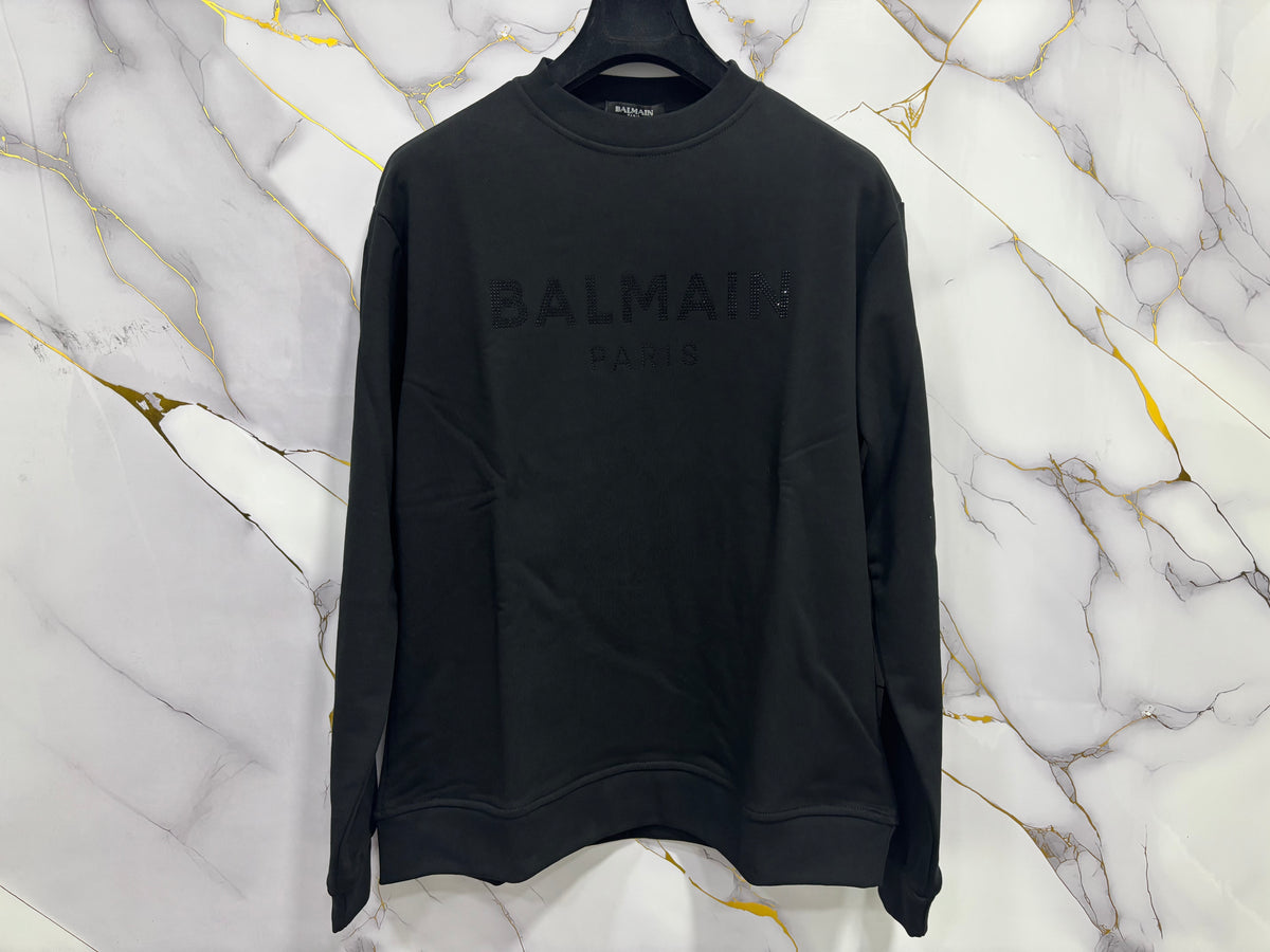 FELPA BALMAIN BRILLANTINATA in black, featuring a sleek design and bold branding on a marble background.